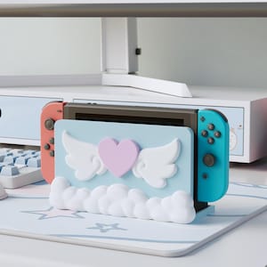 Peut inclure: Support bleu clair pour Nintendo Switch avec un motif de cœur et d'ailes. Le support a une base en forme de nuage et maintient la console Switch avec des manettes corail et turquoise. Le support est sur un tapis à motifs d'étoiles.