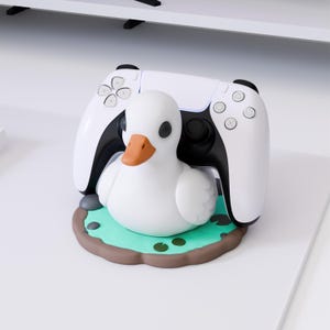 Könnte beinhalten: Ein weißer Controller-Ständer in Entenform mit orangefarbenem Schnabel und schwarzen Augen, der auf einer runden Basis ruht, die wie ein grüner Teich mit braunen Rändern gestaltet ist. Ein schwarz-weißer Videospiel-Controller steht hinter der Ente.