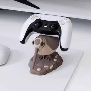 Könnte beinhalten: Ein weiß-schwarzer kabelloser Gamecontroller auf einem braunen Ständer in Form eines Kiwivogels. Der Ständer zeigt einen geformten Kiwivogel mit langem Schnabel, der auf einer strukturierten Basis mit kleinen kieselartigen Details ruht.
