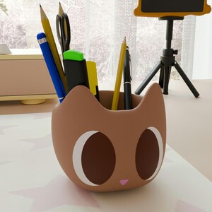 Pode incluir: Um organizador de mesa marrom em forma de gato cheio de canetas, lápis e tesouras. O organizador tem olhos grandes e ovais e um pequeno nariz rosa. Uma caneta azul, um lápis amarelo e uma caneta preta são visíveis.