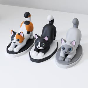May include: Three cat figurines in a stretching pose, each mounted on an oval base. One is white with orange and black patches, one is black with a white chest and face markings, and one is light gray with darker gray ears, face, and tail.