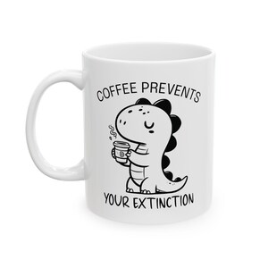 Könnte beinhalten: Weiße Keramik-Kaffeetasse mit einem schwarz-weißen Cartoon-Dinosaurier, der eine Kaffeetasse hält. Auf der Tasse steht in schwarzer Schrift "COFFEE PREVENTS YOUR EXTINCTION". Die Tasse hat einen weißen Henkel.