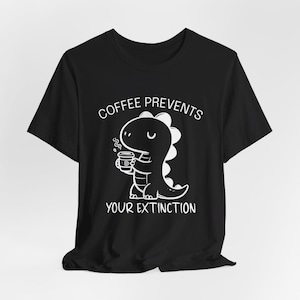 Op de afbeelding: Zwart T-shirt met een witte afbeelding van een dinosaurus die koffie drinkt. De tekst "Coffee Prevents Your Extinction" staat op het T-shirt gedrukt.