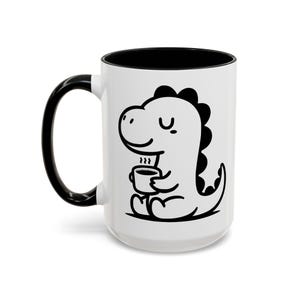 May include: White ceramic mug with a black interior and handle. The mug features a black and white cartoon dinosaur holding a cup of coffee. The dinosaur is smiling, and steam rises from the cup.