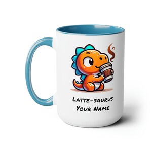 Könnte beinhalten: Eine weiße Keramiktasse mit blauem Rand und Henkel. Die Tasse zeigt eine Cartoon-Illustration eines kleinen orangefarbenen Dinosauriers, der eine Tasse Kaffee hält. Der Text "LATTE-SAURUS YOUR NAME" ist unter dem Dinosaurier aufgedruckt.
