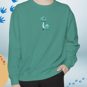 Peut inclure: Un sweat-shirt vert sarcelle avec un motif de dinosaure de dessin animé sur le devant. Le dinosaure est vert avec des accents roses et a une expression amicale.
