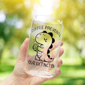 May include: A clear glass can with a black and white illustration of a dinosaur drinking coffee. The text on the can reads "Coffee Prevents Your Extinction."