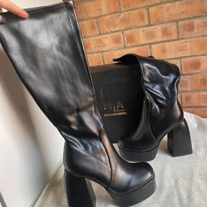 Könnte beinhalten: Schwarze kniehohe Stiefel mit Plateausohlen und Blockabsätzen. Die Stiefel haben eine glänzende Oberfläche und eine runde Spitze. Die Absätze sind dick und quadratisch. Sie werden neben einer schwarzen Box mit dem Text "MIA Shoes" präsentiert.
