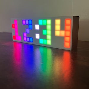 Może przedstawiać: Biały, prostokątny zegar z wyświetlaczem LED w kolorach tęczy, pokazującym godzinę 1:33:34. Zegar stoi na drewnianej powierzchni.