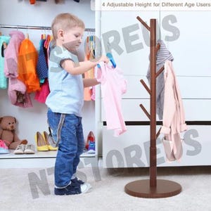 May include: A brown wooden coat rack with four adjustable heights. The rack has six branches for hanging clothes. A young child is standing next to the rack, holding a pink shirt.