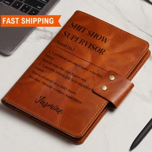 Könnte beinhalten: Ein braunes, ledergebundenes Notizbuch mit dem Text "Shit Show Supervisor" und einer Definition des Begriffs. Das Notizbuch trägt einen personalisierten Namen, "Jasmine", der unten eingraviert ist.