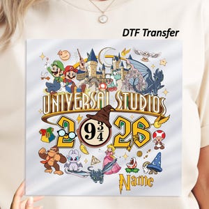 May include: A cream-colored t-shirt featuring a colorful DTF transfer graphic with the text Universal Studios 2026 and Name. The design includes various cartoon characters, a castle, a wizard hat, and the number 9 3/4 in a circular frame.