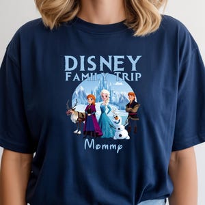 May include: Navy blue t-shirt featuring a graphic of characters from the Disney movie Frozen, including Elsa, Anna, Kristoff, and Olaf, with the text "Disney Family Trip Mommp". The design is set against a snowy mountain backdrop.