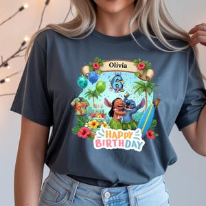 May include: A dark gray t-shirt featuring a colorful graphic of Lilo and Stitch celebrating a birthday. The design includes the name "Olivia", the number 6, tropical flowers, and the words "Happy Birthday".