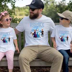 May include: White t-shirts with a Disney Family Trip design featuring characters from Frozen. The shirts are worn by a family of three, sitting on a wooden bench. The text "Disney Family Trip" is printed in blue.