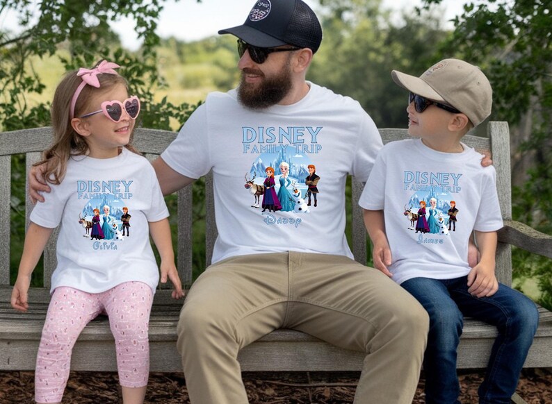 May include: White t-shirts with a Disney Family Trip design featuring characters from Frozen. The shirts are worn by a family of three, sitting on a wooden bench. The text "Disney Family Trip" is printed in blue.