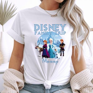 May include: White t-shirt featuring a Disney Family Trip design with characters from the movie Frozen. The design includes Anna, Elsa, Kristoff, Olaf, and Sven, with the word "Mommp" below.