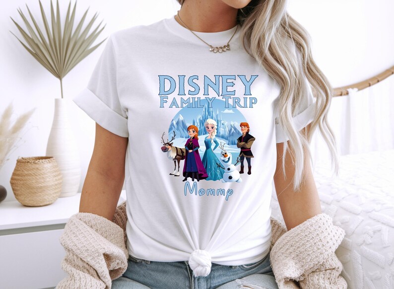 May include: White t-shirt featuring a Disney Family Trip design with characters from the movie Frozen. The design includes Anna, Elsa, Kristoff, Olaf, and Sven, with the word "Mommp" below.