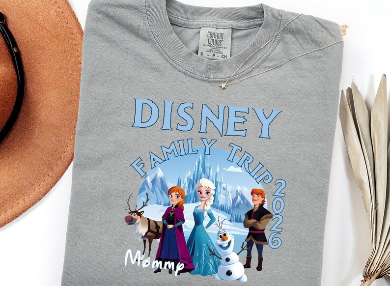 Może przedstawiać: Szara koszulka Comfort Colors z motywem Disney Family Trip z postaciami z filmu Kraina Lodu. Projekt zawiera tekst "Disney Family Trip 2026" i słowo "Mommy". Widoczny jest r&oacute;wnież złoty naszyjnik i brązowy kapelusz.