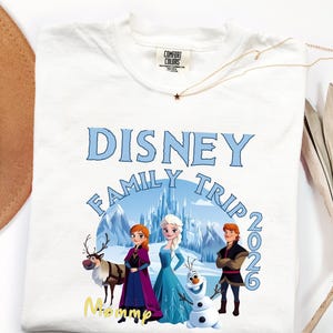 Może przedstawiać: Biała koszulka z nadrukiem Disney Family Trip 2026 z postaciami z filmu Kraina Lodu. Projekt zawiera napis "Disney Family Trip" w kolorze niebieskim, z rokiem 2026. Złoty naszyjnik i kapelusz r&oacute;wnież znajdują się na zdjęciu.