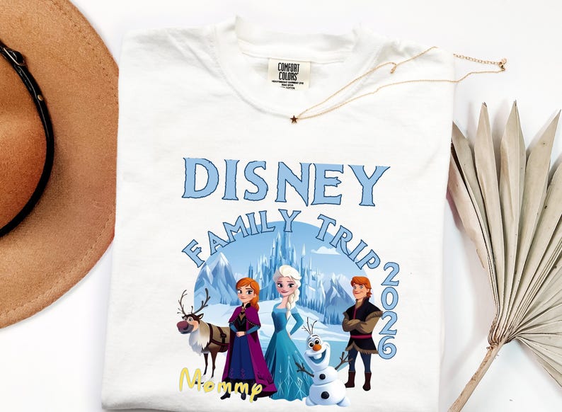 Może przedstawiać: Biała koszulka z nadrukiem Disney Family Trip 2026 z postaciami z filmu Kraina Lodu. Projekt zawiera napis "Disney Family Trip" w kolorze niebieskim, z rokiem 2026. Złoty naszyjnik i kapelusz r&oacute;wnież znajdują się na zdjęciu.