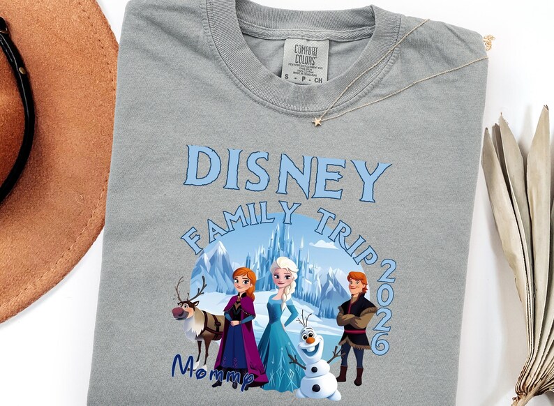 Może przedstawiać: Szara koszulka Comfort Colors z motywem Disney Family Trip. Projekt zawiera napis "Disney Family Trip 2026" oraz grafikę postaci z filmu Kraina Lodu. Widoczny jest r&oacute;wnież złoty naszyjnik z zawieszką w kształcie gwiazdy.