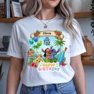 May include: White t-shirt featuring a colorful graphic with the text "Olivia" and "Happy Birthday." The design includes cartoon characters, palm trees, balloons, and a surfboard. The number 6 is also visible in the design.