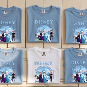 Może przedstawiać: Siedem koszulek, sześć jasnoniebieskich i jedna biała, z nadrukiem Disney Family Trip 2026. Projekt przedstawia postacie z filmu Kraina Lodu. Koszulki są złożone na jasnej drewnianej powierzchni.