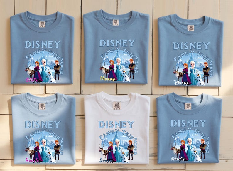Może przedstawiać: Siedem koszulek, sześć jasnoniebieskich i jedna biała, z nadrukiem Disney Family Trip 2026. Projekt przedstawia postacie z filmu Kraina Lodu. Koszulki są złożone na jasnej drewnianej powierzchni.