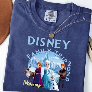 Może przedstawiać: Granatowa koszulka z napisem "DISNEY FAMILY TRIP 2026" w jasnoniebieskim kolorze. Koszulka przedstawia grafikę postaci z filmu Kraina Lodu. Złoty naszyjnik z wisiorkiem jest zarzucony na koszulkę.
