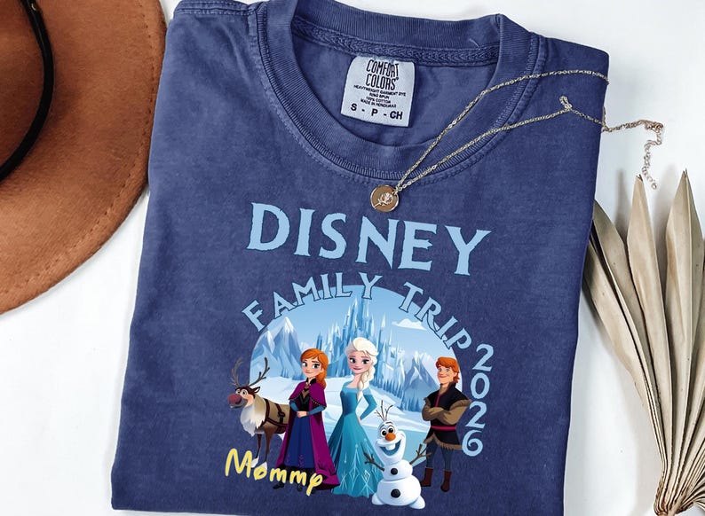 Może przedstawiać: Granatowa koszulka z napisem "DISNEY FAMILY TRIP 2026" w jasnoniebieskim kolorze. Koszulka przedstawia grafikę postaci z filmu Kraina Lodu. Złoty naszyjnik z wisiorkiem jest zarzucony na koszulkę.