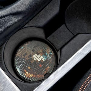May include: A car cup holder with a disco ball coaster. The coaster is round and covered in small, reflective squares. The cup holder is black and sits in a silver-colored trim. The surrounding interior is black leather with orange stitching.