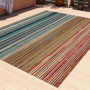May include: A colorful striped outdoor rug with a gradient of blue, green, yellow, orange, and red stripes. The rug is laid out on a wooden deck.
