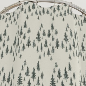 May include: A cream-colored shower curtain with a repeating pattern of dark green pine trees. The trees are stylized and arranged in a wave-like pattern across the fabric. The curtain hangs from a curved, silver-colored shower rod.