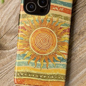 May include: A colorful phone case with a retro design featuring a sunburst in the center. The case has a striped background in shades of orange, yellow, and blue. The case is on a wooden surface.