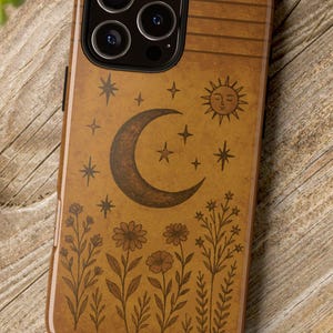 May include: A brown phone case with a celestial design featuring a crescent moon, stars, and a sun. The case also has a floral pattern at the bottom. The phone case is on a wooden surface.