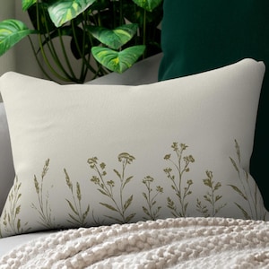 May include: A rectangular cream-colored pillow with a botanical design of wildflowers and grasses in shades of gold. The pillow is placed on a gray sofa with a cream-colored blanket. A green plant and a dark green pillow are in the background.