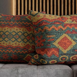 May include: Two square cushions with a colourful geometric pattern in shades of orange, teal, and gold. The cushions are on a grey sofa with a patterned blanket. The cushions have a tribal design.