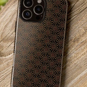 May include: A black smartphone case with a repeating pattern of orange sun-like spirals. The case is on a wooden surface, with a camera and plant in the background. The case has a glossy finish.