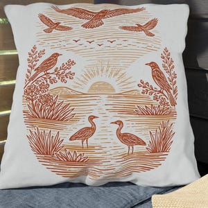 May include: A white square cushion featuring a nature scene in orange and brown. The design includes birds, plants, and a sunset. The cushion is placed on a blue seat.