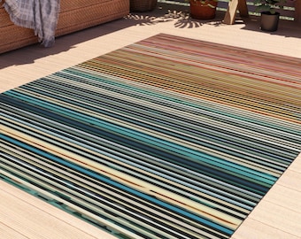 Alfombra para exteriores con rayas en tonos azules y óxido. Esta alfombra presenta rayas que van desde azules y verde azulados intensos hasta naranjas y rojos cálidos. 4 tamaños.