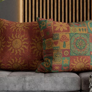 May include: Two decorative throw pillows with a bohemian design. One pillow features a sunburst pattern in shades of orange and yellow. The other pillow has a patchwork design with flowers, mushrooms, and other symbols in orange, green, and yellow.