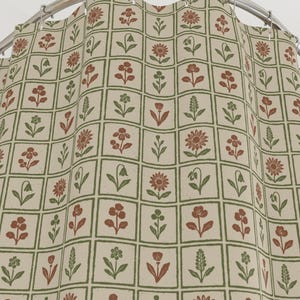 May include: A beige shower curtain with a repeating pattern of red and green floral designs within a grid. The curtain is hanging from a curved, silver-colored shower rod. The floral pattern includes various flower and leaf shapes.