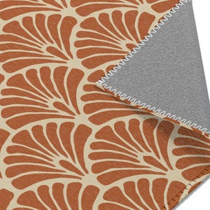 May include: A section of a rug with a repeating fan-shaped pattern in shades of rust and cream. The rug's underside is a gray fabric, and the edges are finished with a white zigzag stitch. The design is a modern take on a classic motif.