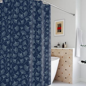 Op de afbeelding: Een donkerblauwe douchegordijn met een wit ginkgo-bladpatroon. Het douchegordijn hangt in een badkamer met een wit bad en een witte betegelde muur.