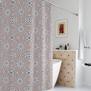 Op de afbeelding: Een douchegordijn met patroon met een herhalend geometrisch ontwerp in tinten wit, grijs en oranje. Het gordijn hangt aan een gebogen, zilverkleurige douchestang. De badkamer heeft een witte badkuip en een neutraal kleurenschema.