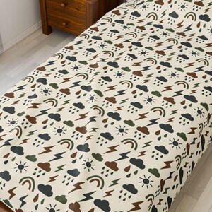 May include: A cream-colored bedspread with a repeating pattern of weather-related icons. The design includes brown, green, and black clouds, rainbows, suns, and lightning bolts. The bedspread is on a wooden bed frame.