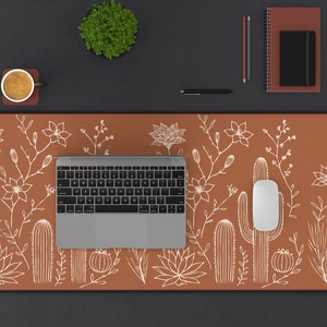 May include: A brown desk mat featuring a white cactus and floral design. A laptop computer, a wireless mouse, and a cup of coffee are on the mat.