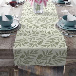 Pode incluir: Um caminho de mesa bege com um padrão repetido de ramos de oliveira em tons de verde. O caminho de mesa está colocado sobre uma mesa de madeira, com talheres que incluem tigelas azuis, pratos brancos e taças de vinho. Um vaso de lírios cor-de-rosa está no centro.