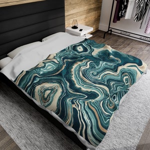 May include: A bed with a white comforter and a teal, beige, and gold patterned blanket. The blanket features a swirling, agate-like design. The bed is against a black headboard and a wooden wall.